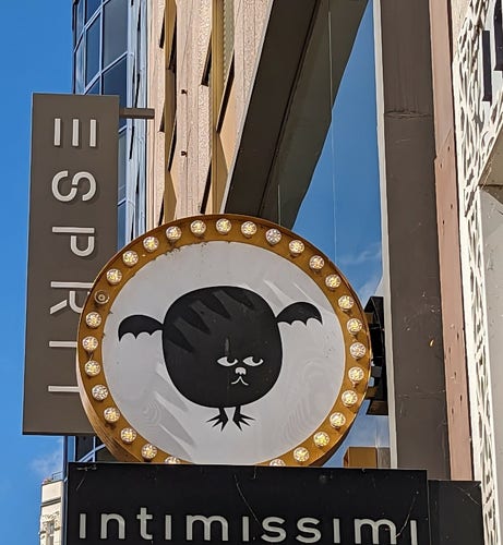 A store sign on the side of a wall. It shows a black round shape with little wings, at the bottom center there's a cartoon face of a cat. The shape also has two small legs that look like they have 3 claws each. The whole figure looks quite dis proportional 