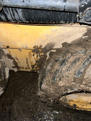 The muddy huge wheel of a skid steer – set in mud – the yellow and black side paneling of the machine – dripping – stained