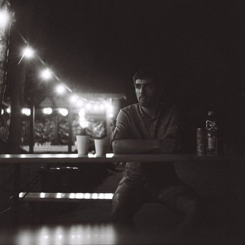 Photo of a man sitting on a bench and looking to the side, lights in the background lightly illuminate his face.
