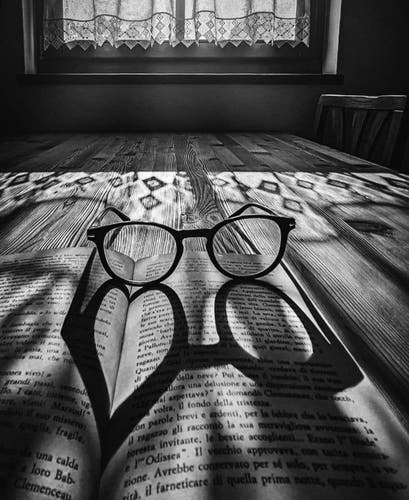 Photography. A black and white photo of a pair of glasses on an open book. The light falls through a window with curtains in the upper center of the photograph. The edge of the curtains casts diamond-shaped shadows across a wooden floor in the room, and the light finally hits the round glasses in the foreground. The interplay of light and shadow creates a dark heart on one of the glasses. A declaration of love to books and reading.