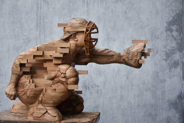 a wooden sculpture of a catcher in baseball, one knee down, his glove outstretched, parts of his body are sculpted realistically and other sections break off into rectangular and cubic shapes 