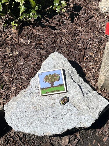 A card depicting a tree with the text "EL ARBOL" and a colorful round sticker are placed on a large triangular rock surrounded by dark mulch.