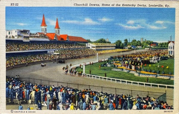 A view of the grandstands, the track, horses racing, and many people cheering under blue skies. 