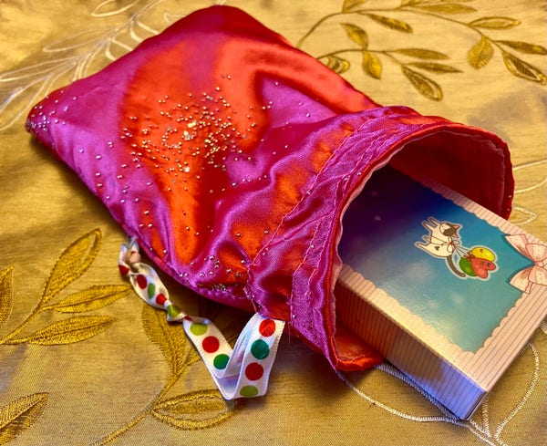 A colorful satin pouch with a deck of cards partially emerging from it, placed on a golden fabric with leaf patterns. The pouch has a polka-dotted ribbon attached to it.