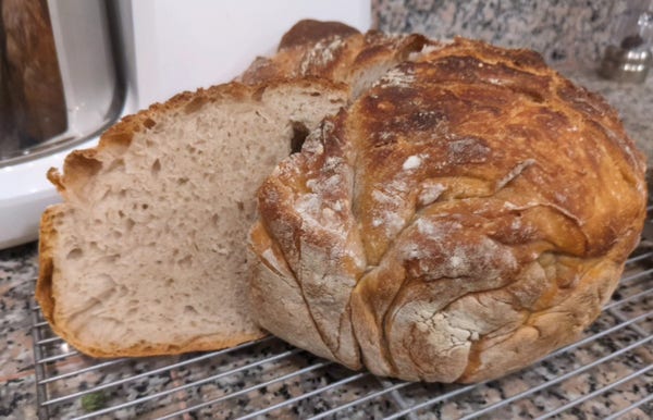 Round loaf of sourdough bread on a cooling rack over a granite counter top.
A slice is pulled aside to show an exemplary distribution of bubbles.
