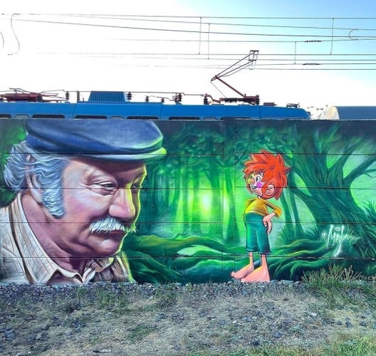 Streetartwall. A large mural with the cartoon character, the goblin "Pumuckl" and the actor Gustl Bayrhammer as "Meister Eder" was sprayed/painted on a long exterior wall. Master Eder is an elderly gentleman with gray hair, a moustache and a flat cap on his head. His head is on the left-hand side of the wall and in front of him stands the red-haired goblin with green, too-short pants, a yellow T-shirt and big feet. He is smiling at him. A green forest has been sprayed on as a background.
Info: Meister Eder and his Pumuckl is a children's book series by Ellis Kaut that was published from 1962 onwards. It is about the childlike goblin Pumuckl, who becomes visible to the Munich master carpenter Franz Eder because he has stuck to his glue pot. According to the "goblin law", Pumuckl must now stay with this human. From 1982, a television series was made with human actors and a Pumuckl as a cartoon character who gets up to all sorts of mischief.