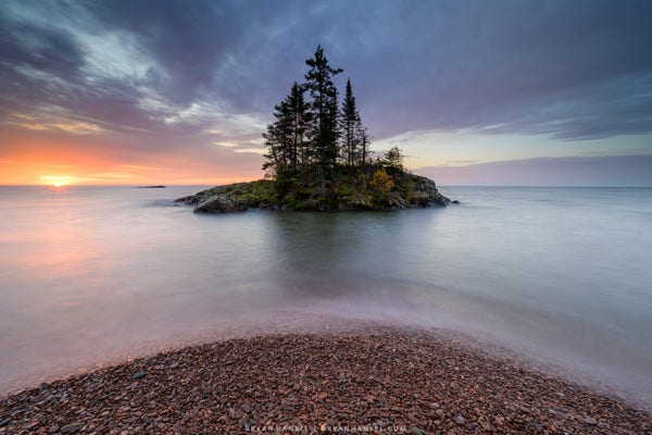 A small round rocky beach in the foreground leads to a smooth Lake Superior and a centered small island with trees and the sun is breaking the horizon just to the left of the island. The photo is full of oranges and purples and blues.