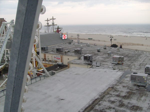 The roof of a building, seen from above. Looking out past the building a beach and large body of water can be seen.

In the foreground a large steel beam with a row of lights on it can be seen, suggestive of some kind of amusement park ride.