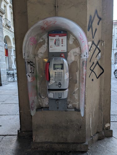 A decaying public phone in Turin, Italy. It is mounted to a large, stained, graffitied marble column in a stone-tiled courtyard. It has a curved transparent cowl, a grey metal body, and a red handset.