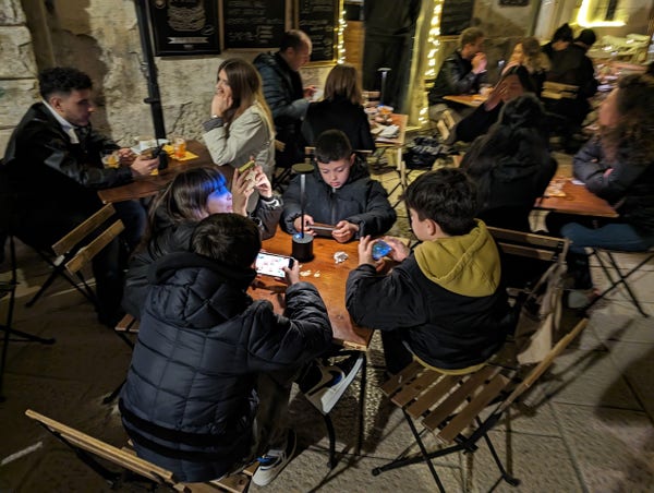 Four kids sittings around a cafe table outdoors at night. They are all holding their phones and gaming together.