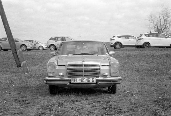 1970s Mercedes car parked on a grass field, with many modern-day cars parked on the road up and behind. Black & white film photo.