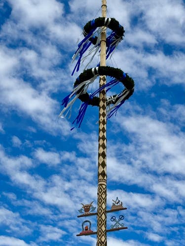 A traditional maypole with blue and white ribbons and wreaths against a cloudy blue sky, featuring symbols such as a pretzel and other icons indicative of local or cultural significance.