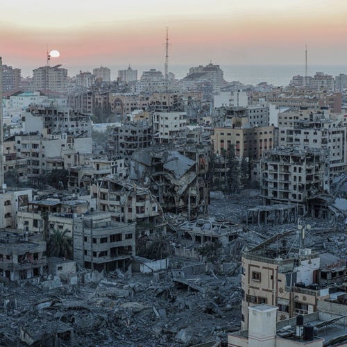 Sunset over a city in Gaza with extensive building destruction and rubble in the foreground, with intact buildings in the background.