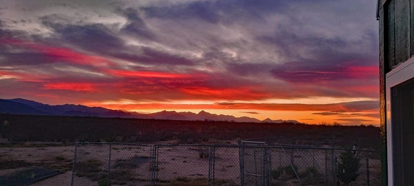 Streaky red and gold sunset across above a brushy, sandy desert. Mountains line the horizon. Chainlink panels are visible in the foreground, some assembled, some still stacked. The edge of a house intrudes on the right side of the picture.