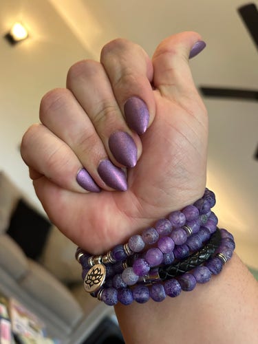 A right hand, palm towards the camera and fingers bent in, showing nails. They are almond shape and have a frosted purple color. On the wrist is a set of mala beads worn as a wrist band. The beads are a similar color as the nails.