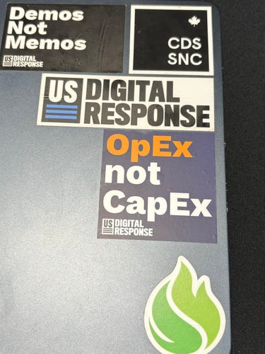 A close-up on a MacBook case, showing several stickers. Centered is one that says “OpEx not CapEx.” Also seen:

“Demos Not Memos”
“US DIGITAL RESPONSE”
“CDS / SNC”