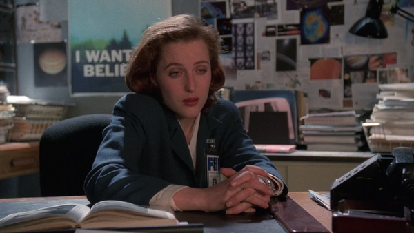 A screencap from X-Files. Scully is sitting at Mulder’s desk, with the “I WANT TO BELIEVE” poster behind her, with an exasperated expression on her face.