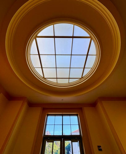 A wide-angle photo of a few magnificent windows taken in a wing at Doe Library on the UC Berkeley campus. We are looking at a huge circular dome window in the center top, below that is a rectangular window above a glass door. Everything is centered and very geometrically aligned with gorgeous lush warm luminous colors in the walls and moulding around the blue windows looking out at other regal campus buildings. Personally, it’s giving me big Bespin Cloud City from Star Wars vibes. I can almost picture Lando Calrissian standing just off-camera with Chewbacca wearing C3-PO on his back to his side. 