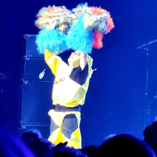 Mark Mothersbaugh, in a yellow vinyl jumpsuit, shaking multicolor pom poms above his head.