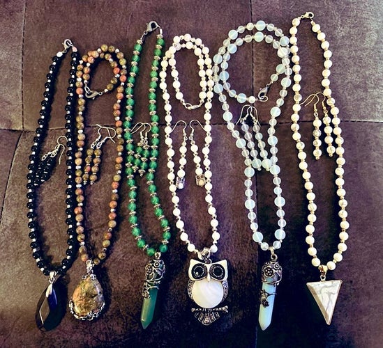 Six necklace and earring sets: black Jasper with teardrop pendant, Unakite with teardrop pendant and flower accent, aventurine with crystal pendant with flowers, white shell with owl pendant, moonstone with crystal pendant and flowers, and white Howlite with triangle pendant 