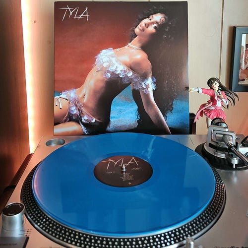 A turqoise vinyl record sits on a turntable. Behind the turntable, a vinyl album outer sleeve is displayed. The front cover shows Tyla sitting on blue sand. She is leaning back with her hands on the ground behind her, as she looks at the camera.