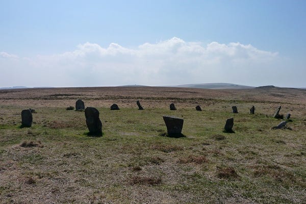 A wide circle of granite stones, standing on an open, grassy moor. The stones appear almost black against the green of the moor. Higher hills rise on the distant skyline, with a rocky tor prominent on the right. The sky is clear blue with some hazy cloud.