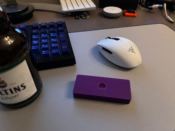 A computer desk with a mechanical keyboard, a white gaming mouse, a bottle of beer, and a purple object, with a phone charger and other items in the background.