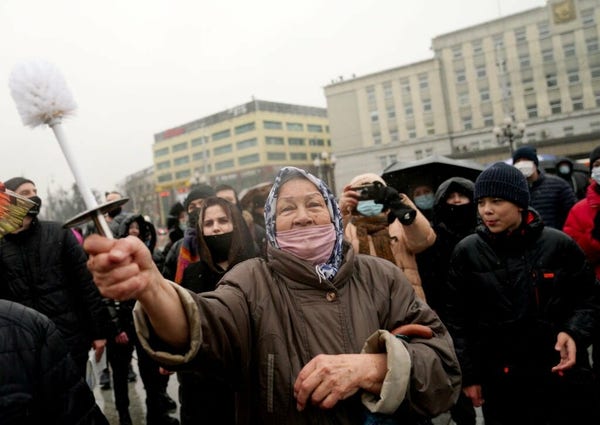 Foto di una protesta di piazza a Mosca, in primo piano una signora anziana con la bocca coperta e un fazzoletto in testa brandisce uno scopino del cesso per protestare contro Putin