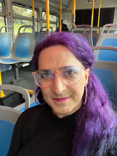Selfie of Nora on a bus. She’s got large purple hoop earrings on which match her long purple hair, and she is looking at the camera with a smirk. She thinks herself rather cute. Do not disabuse her of this notion, for it is a pleasant fantasy. 