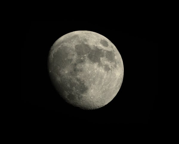 90% illuminated disc of moon against black, starless background.
Shadow line on far left of the moon.