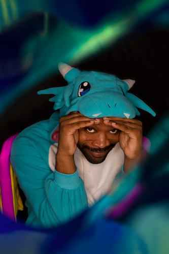 Synxiec is in a dragon onesie. The onesie is a light tone of blue with pink and yellow wings. The hood of the onesie has eyes, teeth, and horns similar to a small dragon.

Synxiec peeks from under his hands while smiling at the camera. An item wrapped around the camera lends makes a multicolored blur at the edges of the photo.
