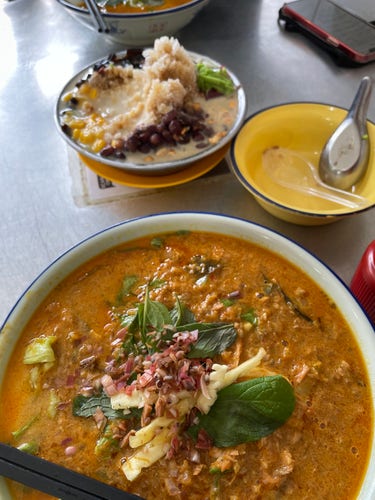 A large bowl of Laksa Siam, a rice noodle dish with thick and creamy fish broth that includes coconut milk and lots of shredded fish meat. There’s a shaved ice dessert in the background.