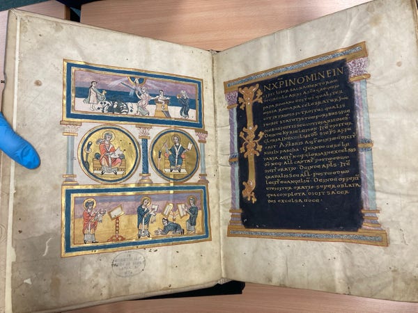 An open manuscript book with ornate illustrations and text, some pages showing gold leaf detailing. A person's hand in a blue glove points to the text.