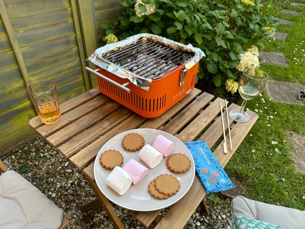 Photograph of a small table top bbq in a garden setting. On the table is a plate of biscuits and marshmallows, a bar of chocolate, skewers, and drinks. 