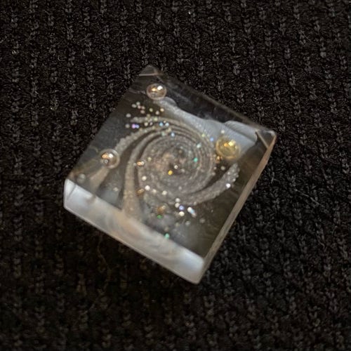 A clear resin square about the size of a chiclet, with a pearlescent white swirl like a multi-armed galaxy, some clusters of holographic glitter stars, and some gold and silver nail art spheres as suns/moons/planets