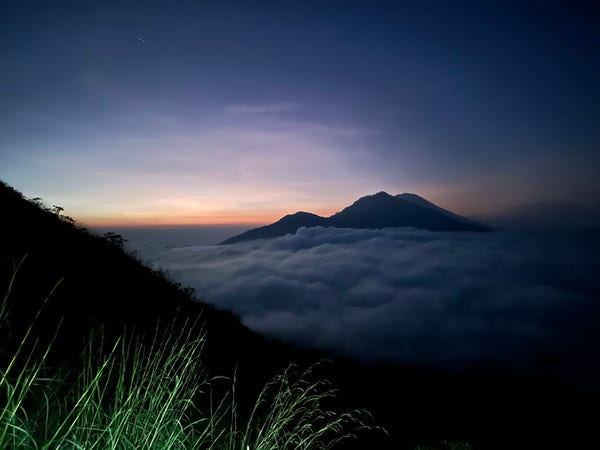 The Gunung Agung volcano clouded in skies. The sun is starting to rise behind it, in red, orange and purple tints. 