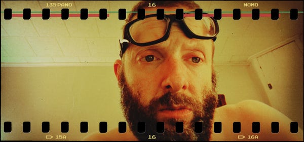 yellow self portrait with strap on glasses pushed up to forehead – forehead wrinkles – red eyes – beard – all in an empty room