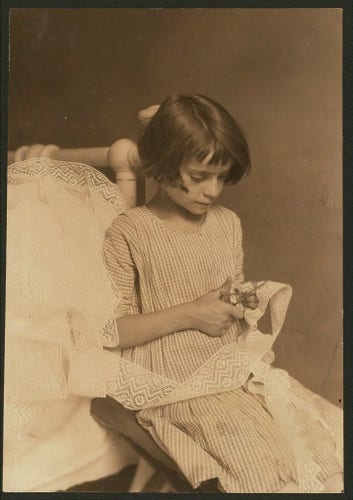  The image is a black and white photograph featuring a young girl seated on what appears to be a sofa or chair. She is wearing a striped dress with puff sleeves, which suggests a style from an earlier time period, possibly the late 19th or early 20th century. The girl has dark hair and is looking down at an item she holds in her hands.

The setting seems to be indoors, perhaps a living room or parlor, indicated by the upholstered furniture behind her. The lighting is soft, casting gentle shadows on her face and dress, which lends a quiet, introspective mood to the image.

There is no visible text in the photograph, and it does not provide any context or information about the girl's identity, location, or the nature of the item she holds. The image has a sense of stillness and timelessness due to its monochromatic quality and the subject's contemplative demeanor.