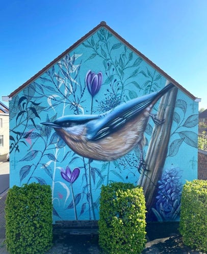 Streetartwall. A beautiful blue nuthatch has been sprayed/painted on the outside wall of a two-story house. The background is a floral scene in blue, with white and green/blue ferns, blue flowers and purple lilacs. The huge bird with blue/white plumage sits diagonally on a broad branch and spreads harmony. (In front of the mural are three green ponds, above which lies a blue, cloudless sky)