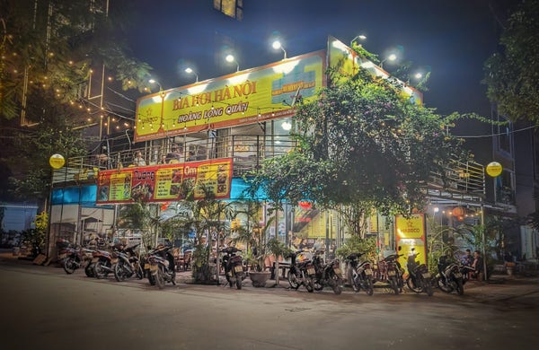 An evening photo of a beer joint on the outskirts of Hanoi. It is a modest, open-air, two-storey structure on a corner beside a large, flowering bougainvillea. The restaurant's brightly lit signs are mostly gold with red lettering, the color scheme of the beer they serve (Bia Hơi Hà Nội). Ground level signs announce menus of hotpot, grilled items, and braised meats. Parked motorbikes form an almost unbroken perimeter on the pavement around the entrance.