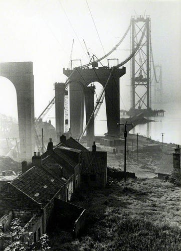 Black and white photo of the Forth Bridge in Edinburgh being constructed. It is a suspension bridge. At the time of this photo, it appears that most of the construction is complete, except for the attaching of the road segments. It's foggy, and in the foreground are some tumbledown-looking old stone houses.