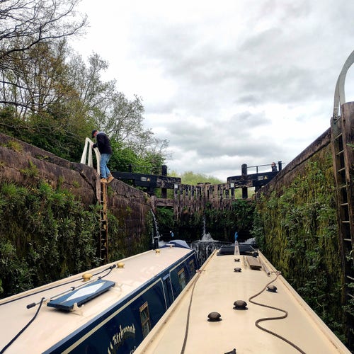 Two narrowboats in a lock that's covered in aquatic plants and weeds. A boater is descending the ladder back to his boat, after helping with the lower gates. At the upper gate another crew member is using a windlass to wind open a paddle and fill the lock chamber with water from the pond above the lock.