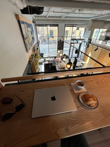 A photo of a trendy Oslo coffee shop, taken from a mezzanine that overlooks the shop. People buy coffee down below. A laptop and coffee and croissant sit on the ledge of the mezzanine. 