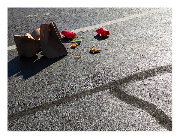 night, lit by security light. litter dumped by a customer in a parking spot at mcdonalds. the asphalt is patched with tar, and there is one white line. two crumpled bags, assorted fries and nuggets a green-labeled container of sauce and two red french fries sleeves.