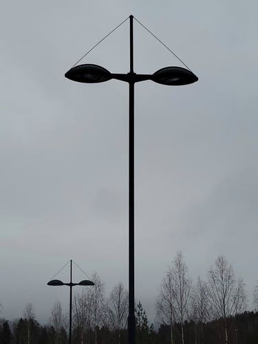 Two black streetlights, both with two lights with a wire attached to them and the end of the tip of the pole, making a triangle. The lights stand in front of a row of treetops against a grey sky. The picture is naturally monochrome