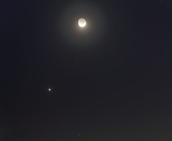 A night sky. A the top, the lit part of the crescent moon is over-exposed and drowned in glare, while the unlit part is quite brightly lit from earth shine. A foggy halo surround the moon. The next brightest thing, below and to the left of the moon is Jupiter, also over-exposed, with bright dots of Callisto and Ganymede almost lost in the glare. Almost directly above Jupiter is a fairly bright bluish "star" which is Uranus. At the bottom center, the comet is a greenish smudge with a faint tail pointing up.