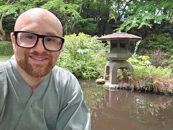 A selfie of me in front of a stone lantern that's on the edge of a pond in a Japanese garden. Under some trees there's small waterfall streaming down strategically placed rocks in the background, and a few koi are barely visible in the tea-colored water. I'm a bald, middle-aged white man with a red beard flecked with white wearing thick black glasses wearing a light green yukata