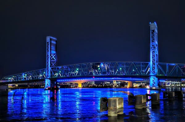 From a wet walkway along the Saint Johns River in downtown, a late night view across the water where both the Main Street & Acosta bridges are illuminated in blue and reflecting upon the calm water below against the backdrop of a colorful skyline.