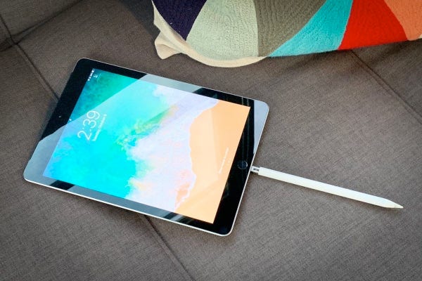 An iPad with a colorful screen display lying on a grey sofa next to a multi-colored throw pillow. An Apple Pencil is sticking out of the iPad’s Lightening port.