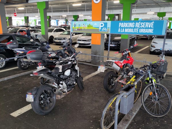 Une photo du parking vélo : 4 motos occupent la majeure partie de l'espace. Trois vélos sont accrochés à la barrière qui délimite le parking. Les seules accroches disponibles pour les vélos sont des pinces-roues...